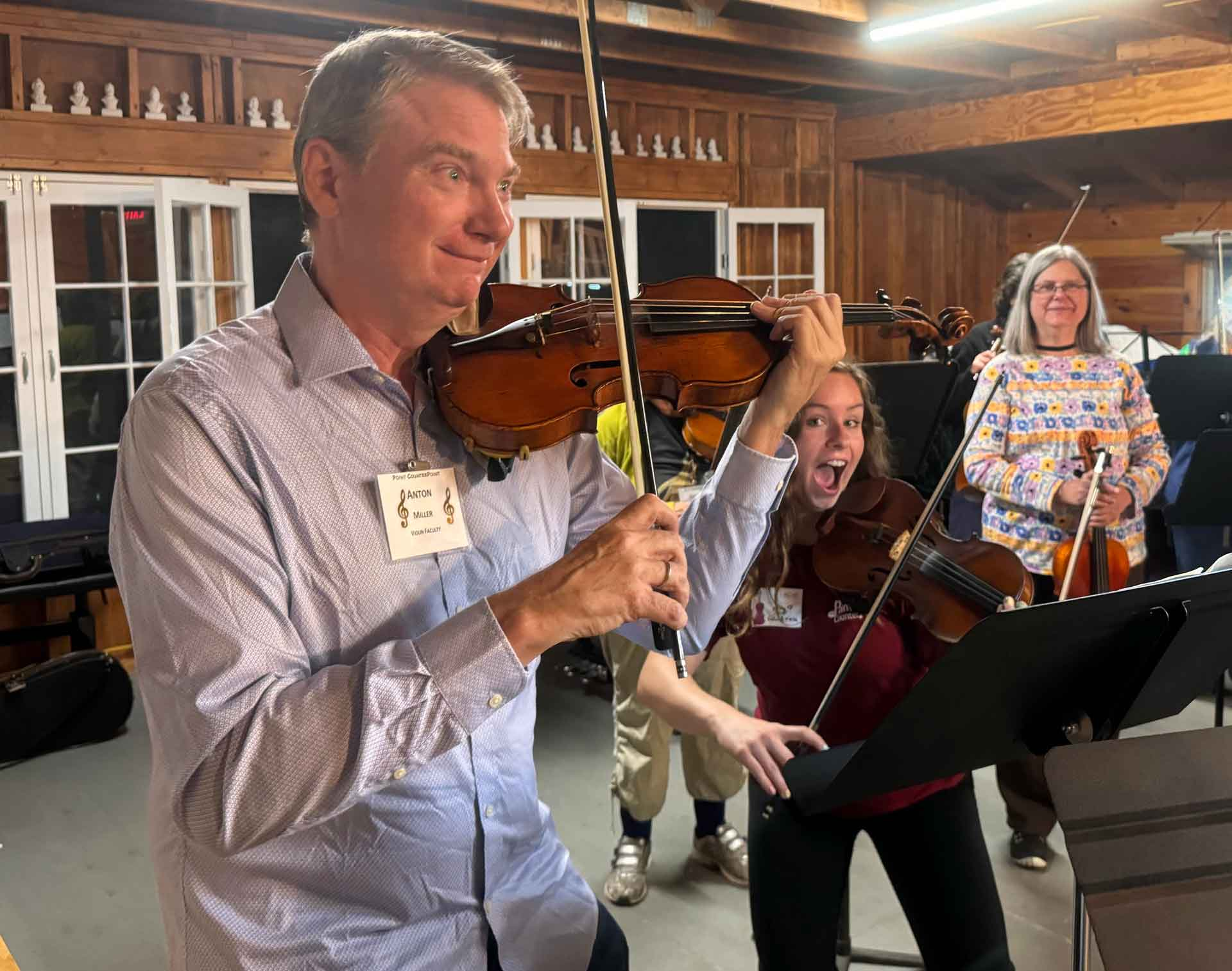 Point CounterPoint Three adults play violins indoors at Point CounterPoint; the man in front smiles while the woman behind him makes a surprised face. Sheet music and wooden walls are visible in the background.