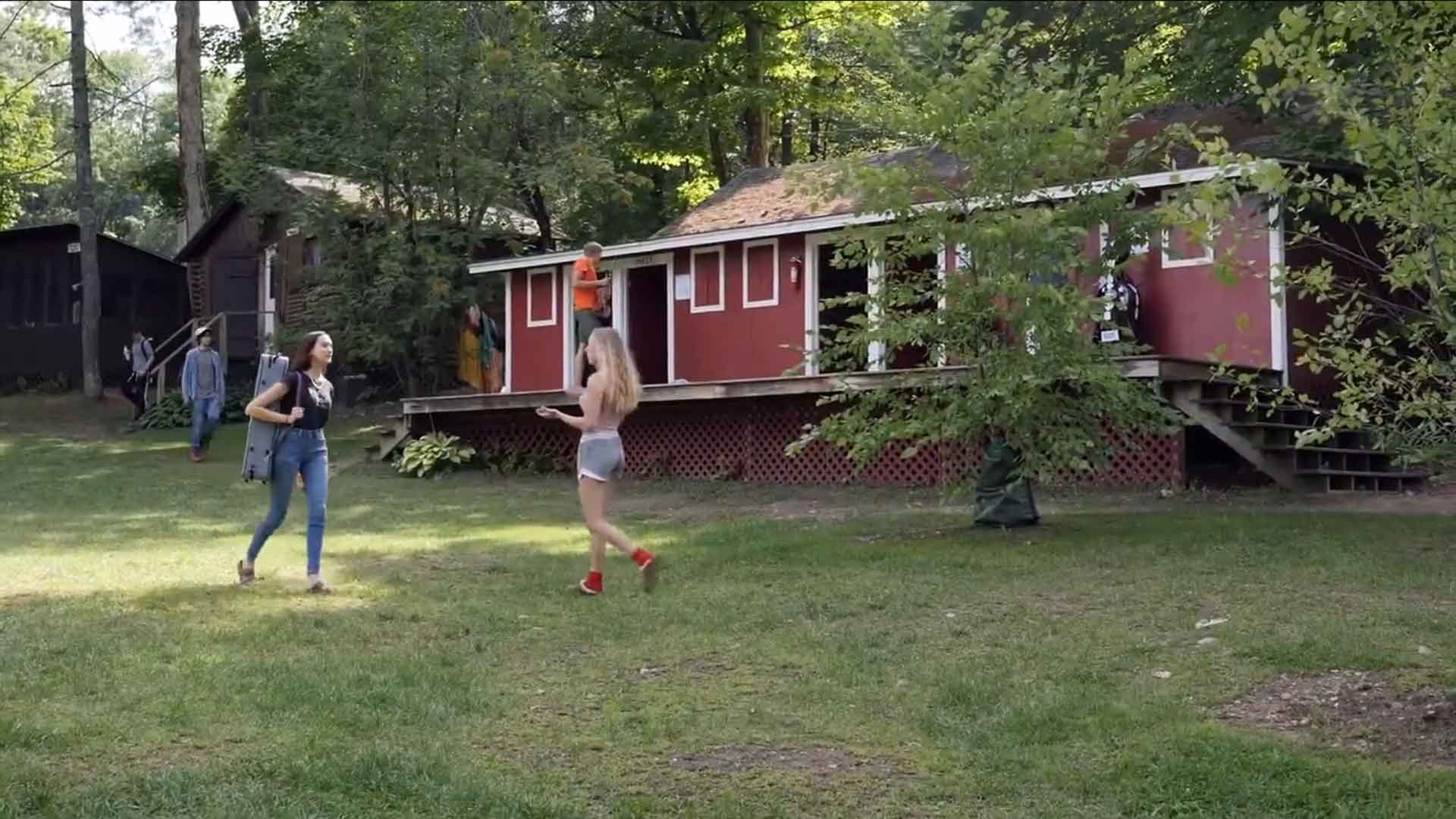 Coming to Camp! Three people are outside near a red cabin surrounded by trees. One person walks across the grass while two others stand and talk in the yard, enjoying Youth Activities designed to promote connection and fun in a natural setting.