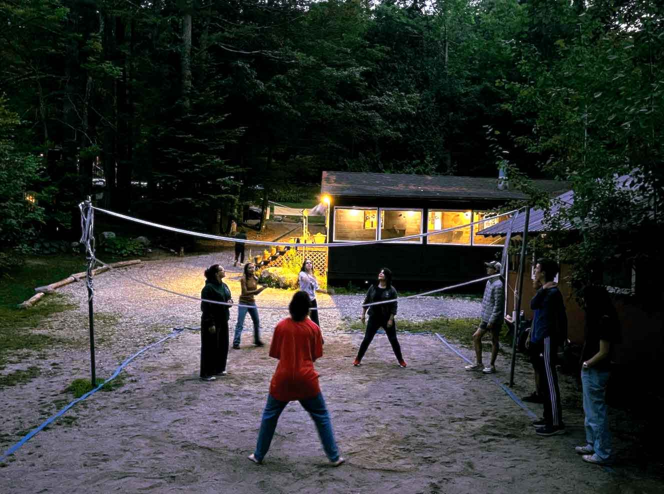 Point CounterPoint A group of people play volleyball on a sandy court outdoors near a cabin at dusk, surrounded by trees, as the friendly match sparks Point CounterPoint debates over each call.