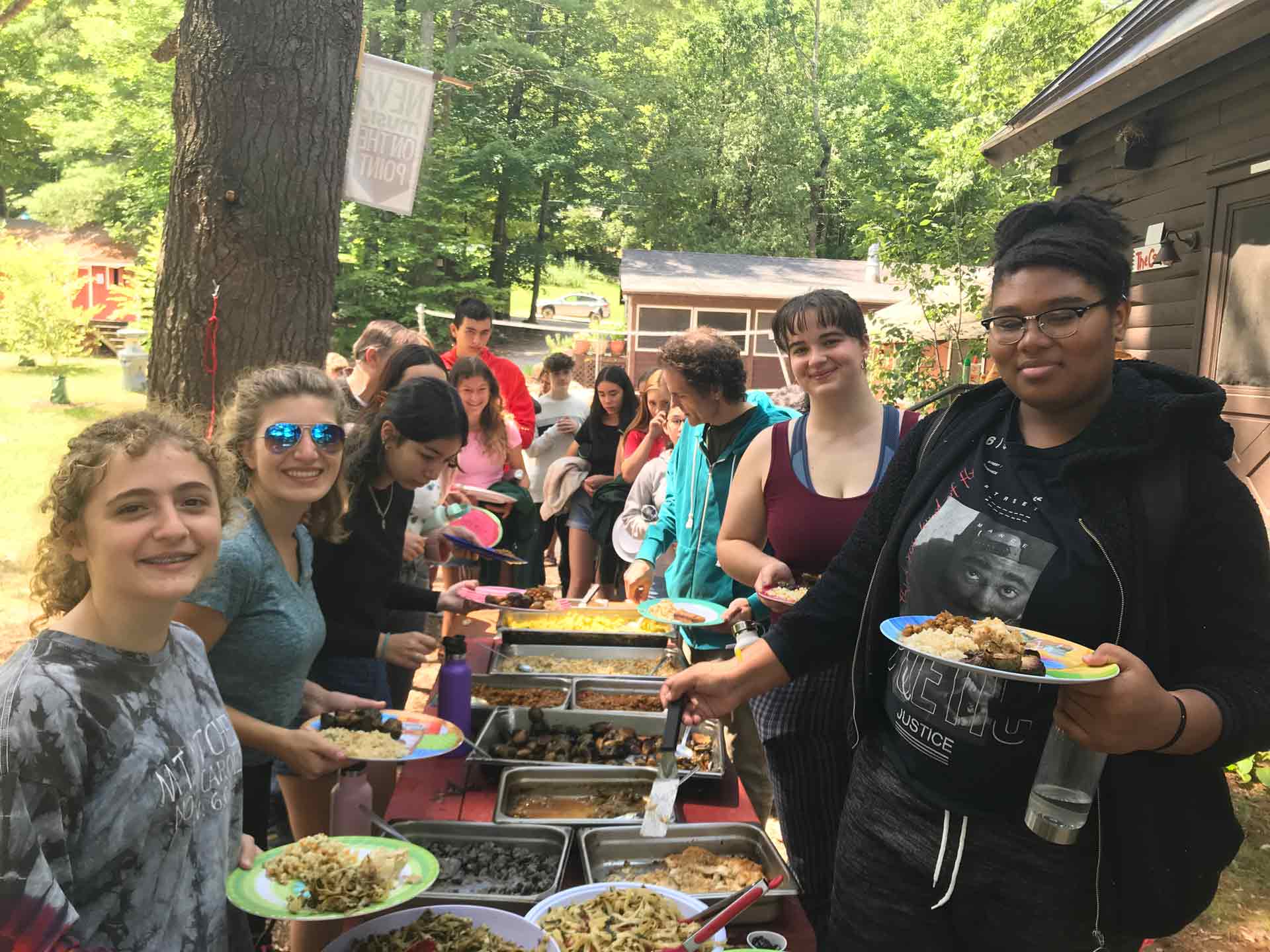 Coming to Camp! A group of people stands in line outdoors at camp, serving themselves food from a long buffet table filled with various dishes, surrounded by trees and wooden buildings.