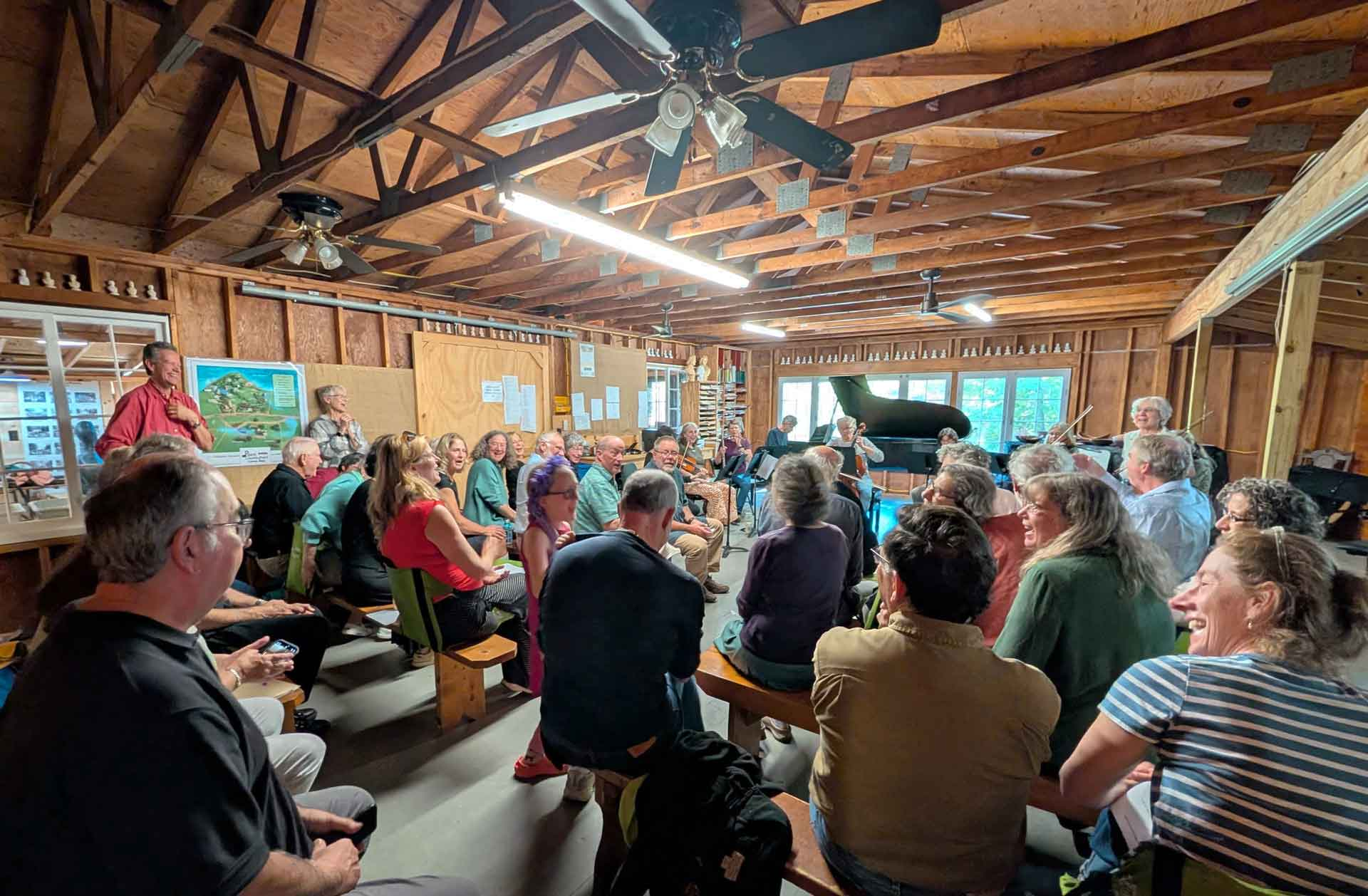 Point CounterPoint A group of people sit on benches in a rustic wooden room, attentively listening to a Point CounterPoint performance by a person playing a grand piano at the front.
