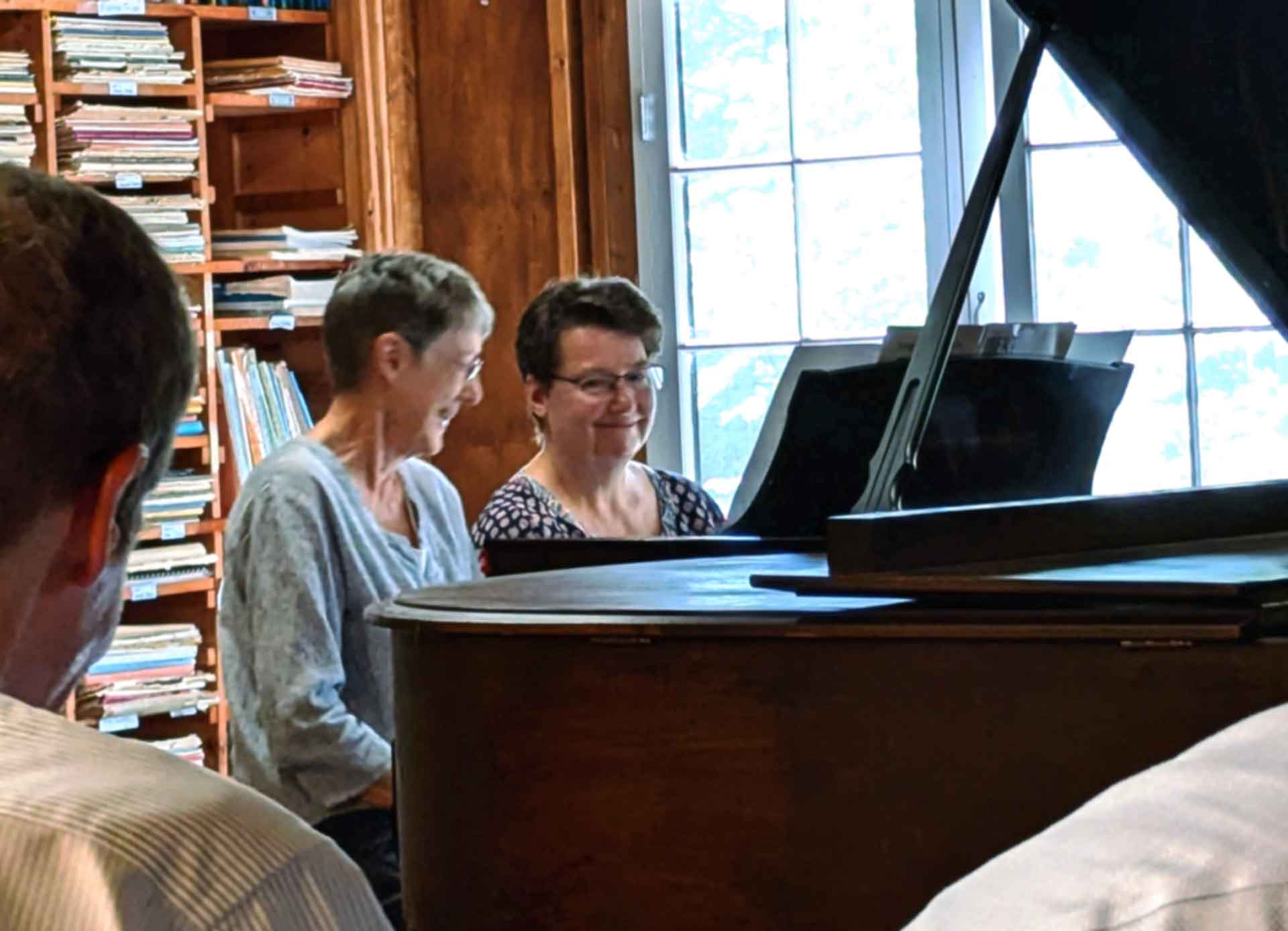 Adult Programs Two women sit together at a grand piano in a classic Point CounterPoint moment—one playing while the other watches and smiles, with bookshelves and a window in the background.