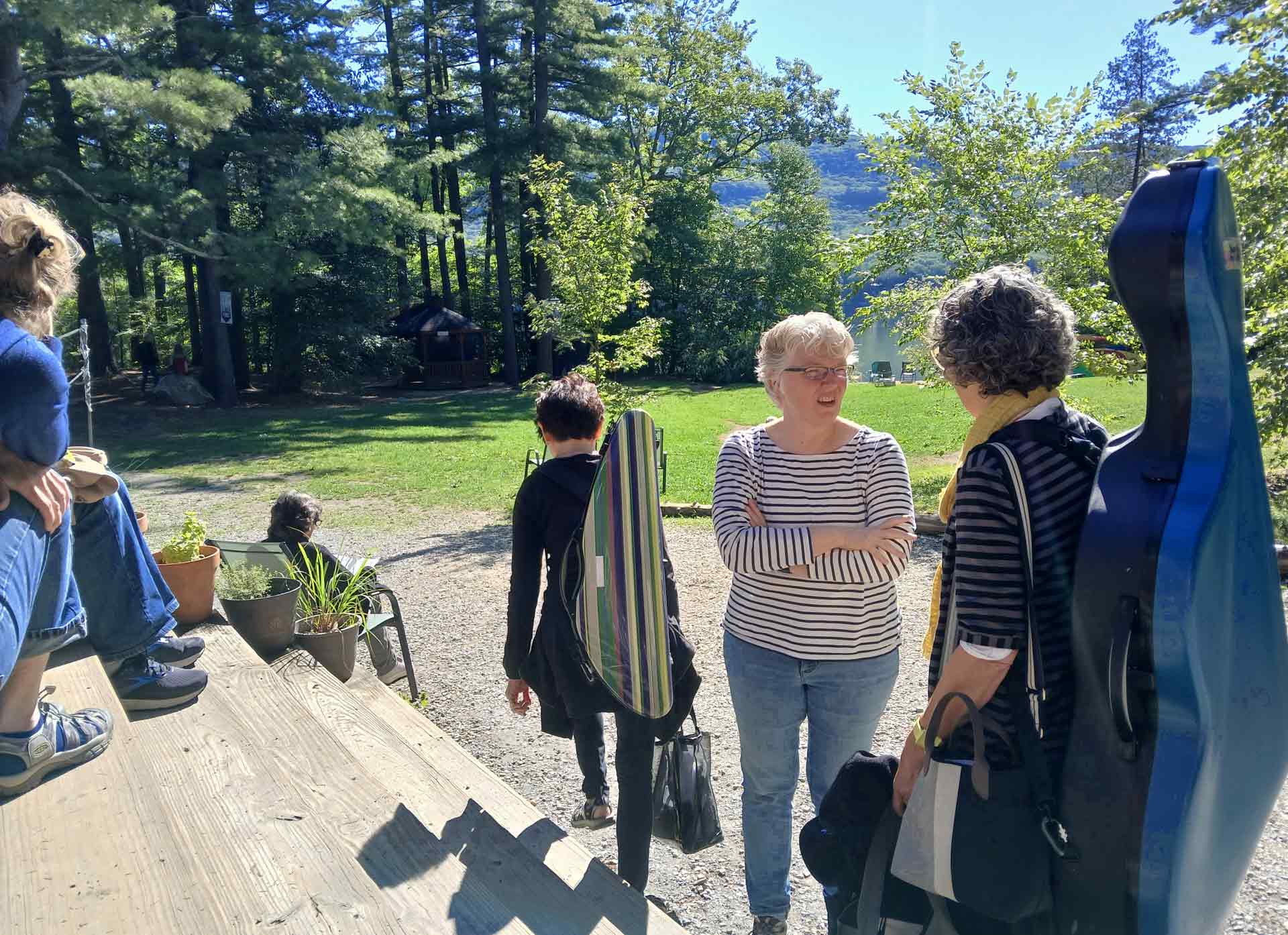 Adult Programs A group of people involved in Adult Programs stand and sit outdoors near wooden steps, talking and holding instrument cases, with trees and sunlight in the background.