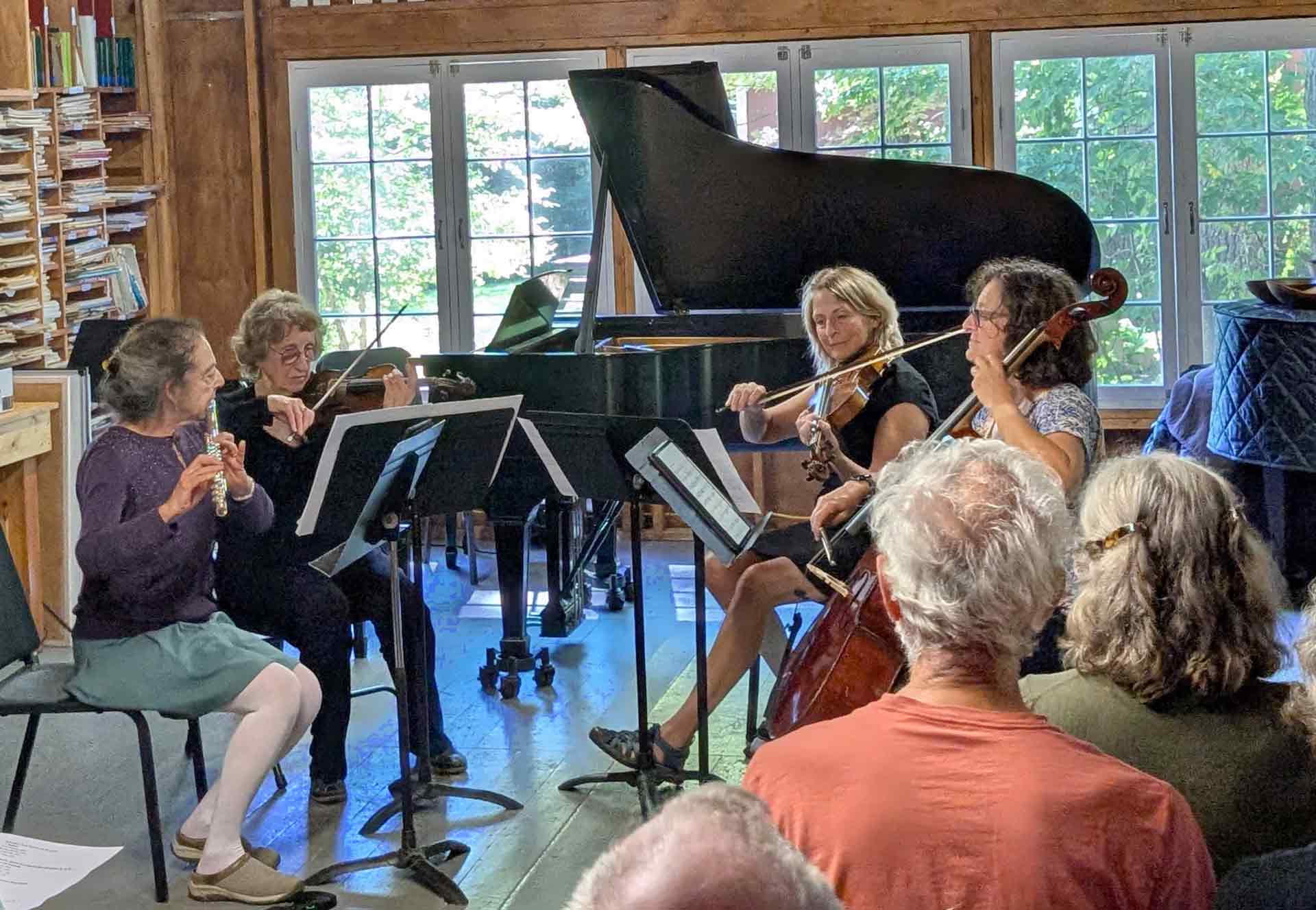 Adult Programs Four musicians perform indoors with string instruments and a flute, seated in front of music stands for an engaging Adult Programs event, while an audience watches; a grand piano is in the background.