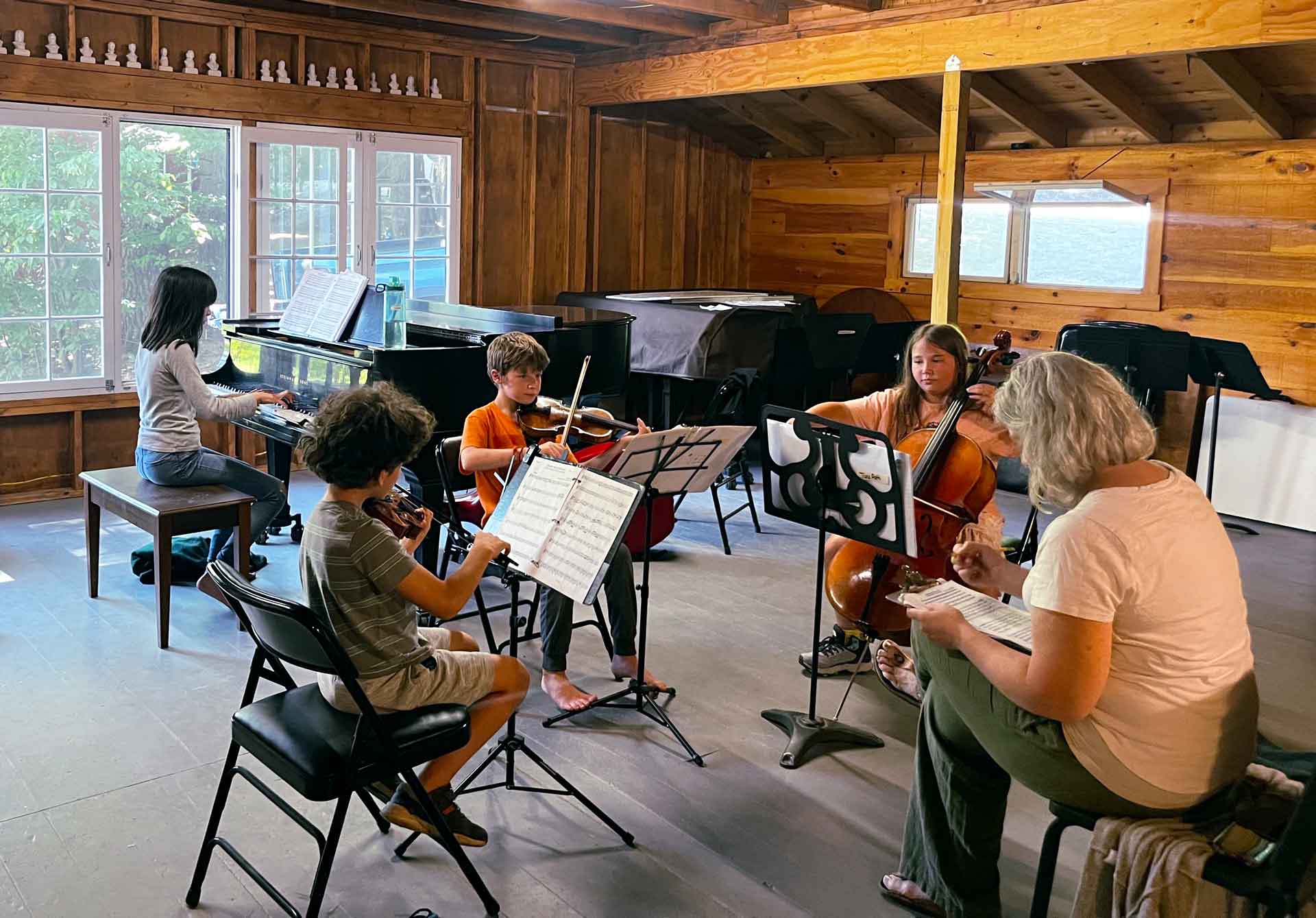 Youth Programs Four children rehearse with string instruments and piano in a wood-paneled room, embodying the spirit of Point CounterPoint, while an adult sits nearby taking notes.