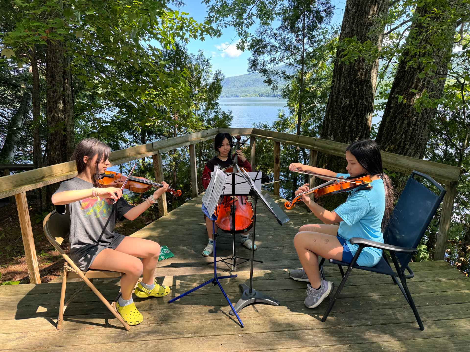 Youth Programs Three girls from local youth programs play string instruments—two on violins, one on cello—outdoors on a wooden deck surrounded by trees, with a lake visible in the background.