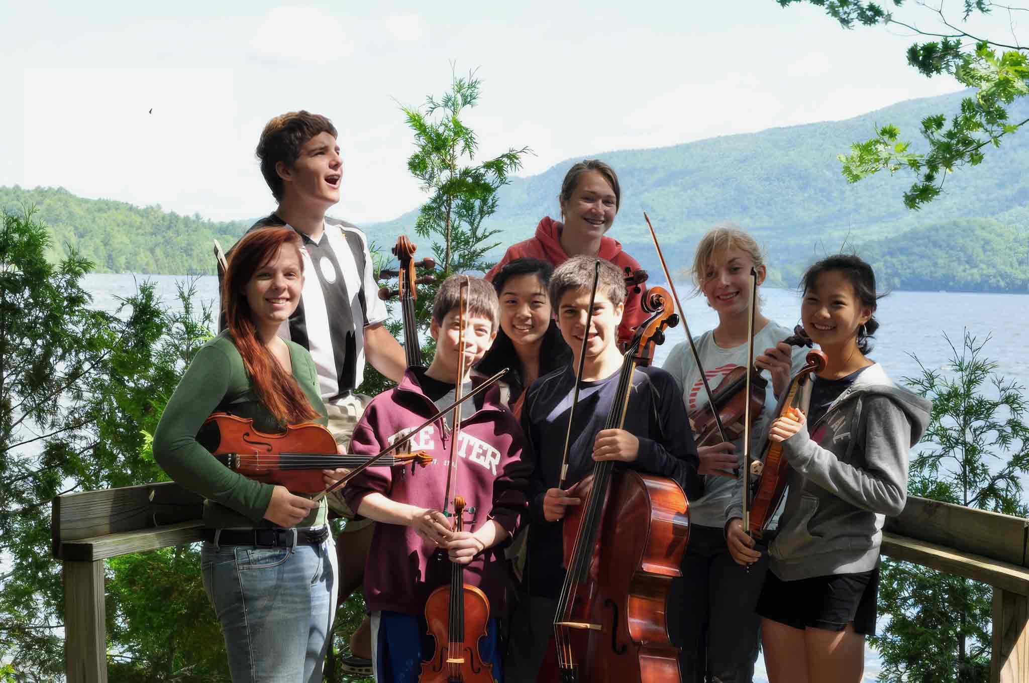 Point CounterPoint A group of seven teenagers and one adult pose outdoors with string instruments, smiling, with a lake and mountains in the background, perfect for showcasing on your main page.
