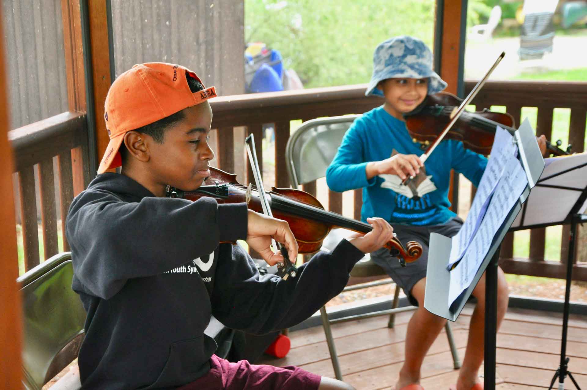 Youth Programs Two children sit on chairs under a wooden gazebo, engaging in youth activities as they play violins and read sheet music on stands. One wears an orange cap, the other a blue bucket hat.