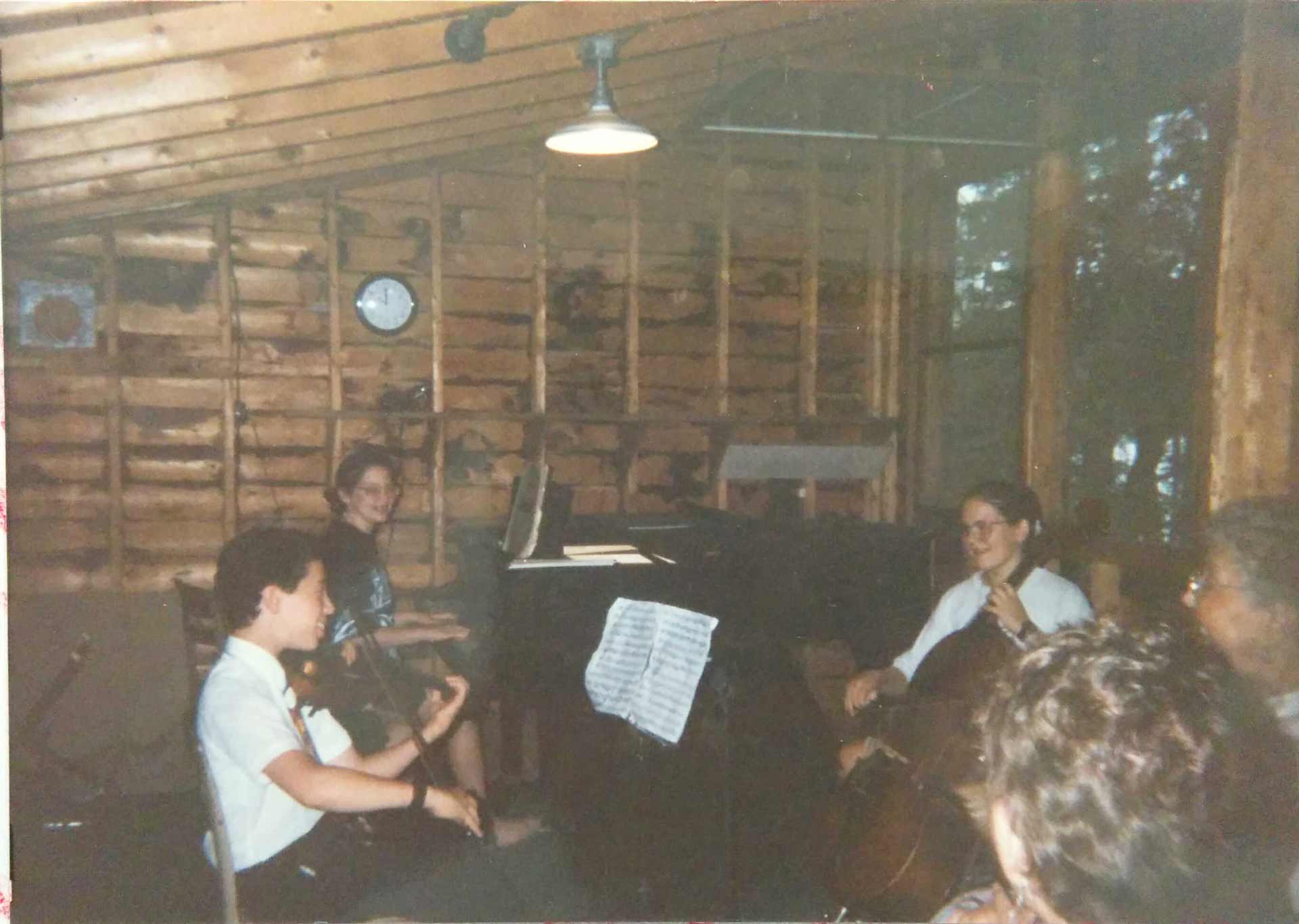 Point CounterPoint History A group of people play string instruments and piano in a rustic wooden room with exposed walls and ceiling, evoking the rich history of chamber music. Sheet music is visible on a stand by the piano—a scene reminiscent of Point CounterPoint gatherings.