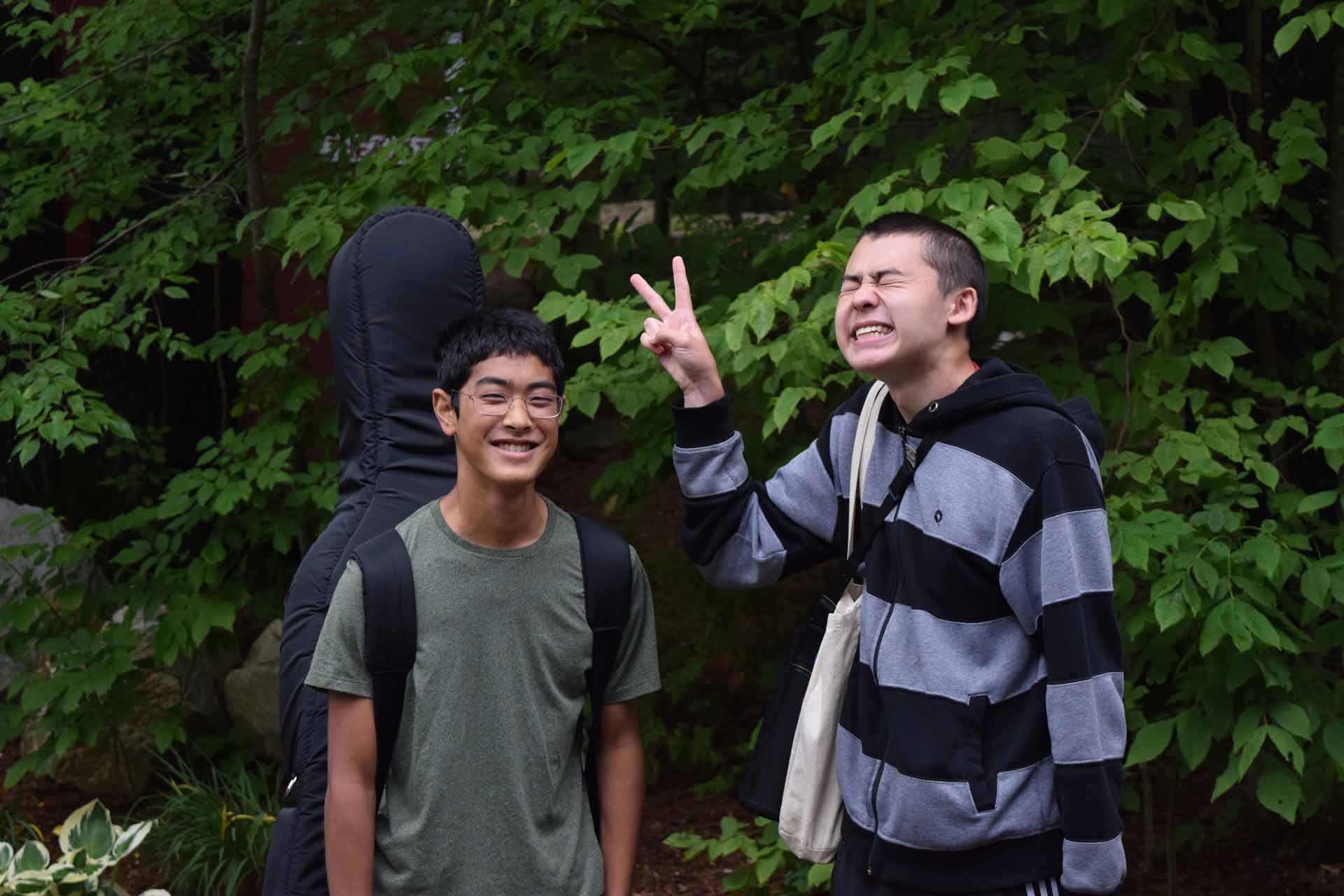 Play it Forward Campaign Two young men stand outdoors; one smiles while the other makes a peace sign gesture and grins enthusiastically, embodying the fun spirit of the Play it Forward Campaign. Green foliage fills the background.
