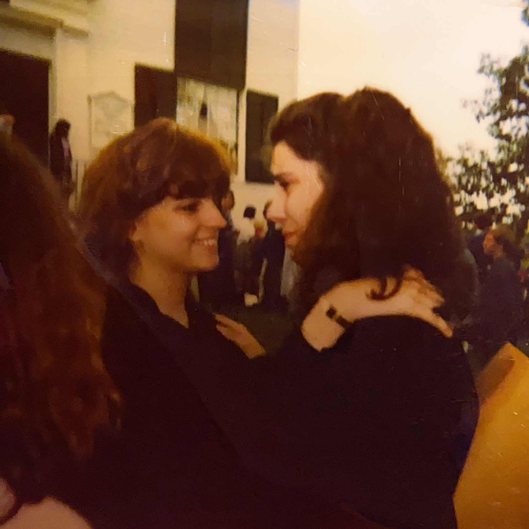 Point CounterPoint History Two women face each other and embrace outdoors, one smiling and the other appearing emotional, capturing a Point CounterPoint History moment with people and a building in the background.