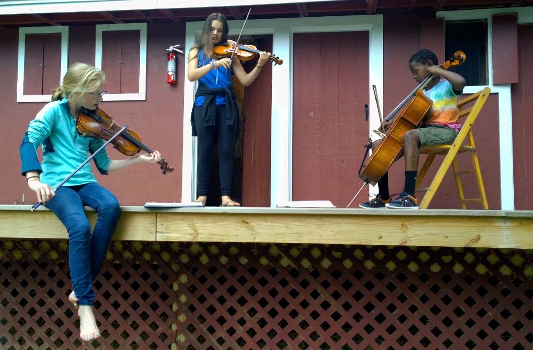 About Point CounterPoint Three young musicians, part of Point CounterPoint, play string instruments—two violins and a cello—on a wooden porch in front of a red building.
