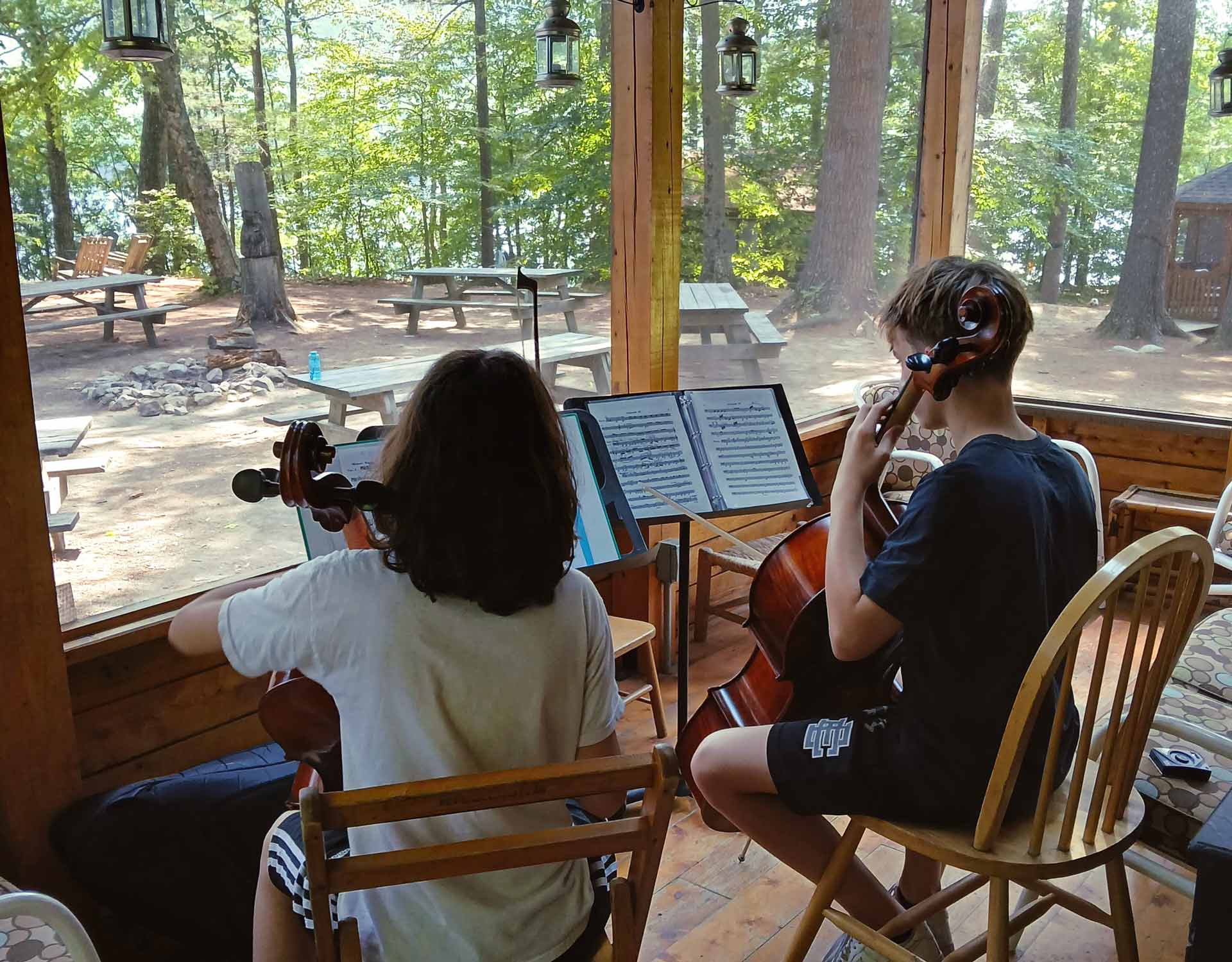 Play it Forward Campaign Two children play cellos indoors near large windows with sheet music, as a wooded area and picnic tables are visible outside—a perfect scene to inspire the Play it Forward Campaign.