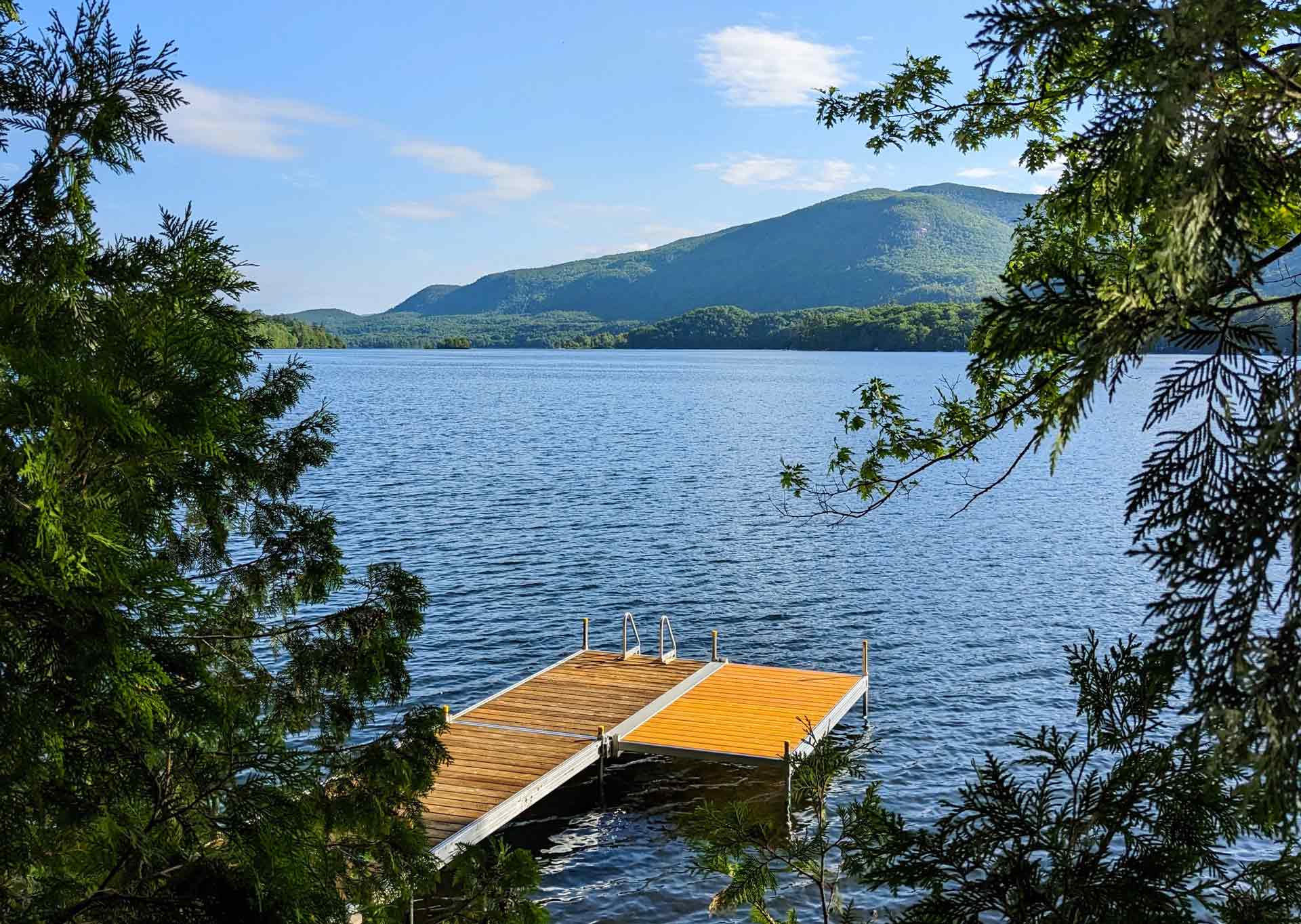About Point CounterPoint A wooden dock extends into a calm lake, surrounded by trees and watched over by Point CounterPoint’s green mountain in the background, all beneath a partly cloudy blue sky.