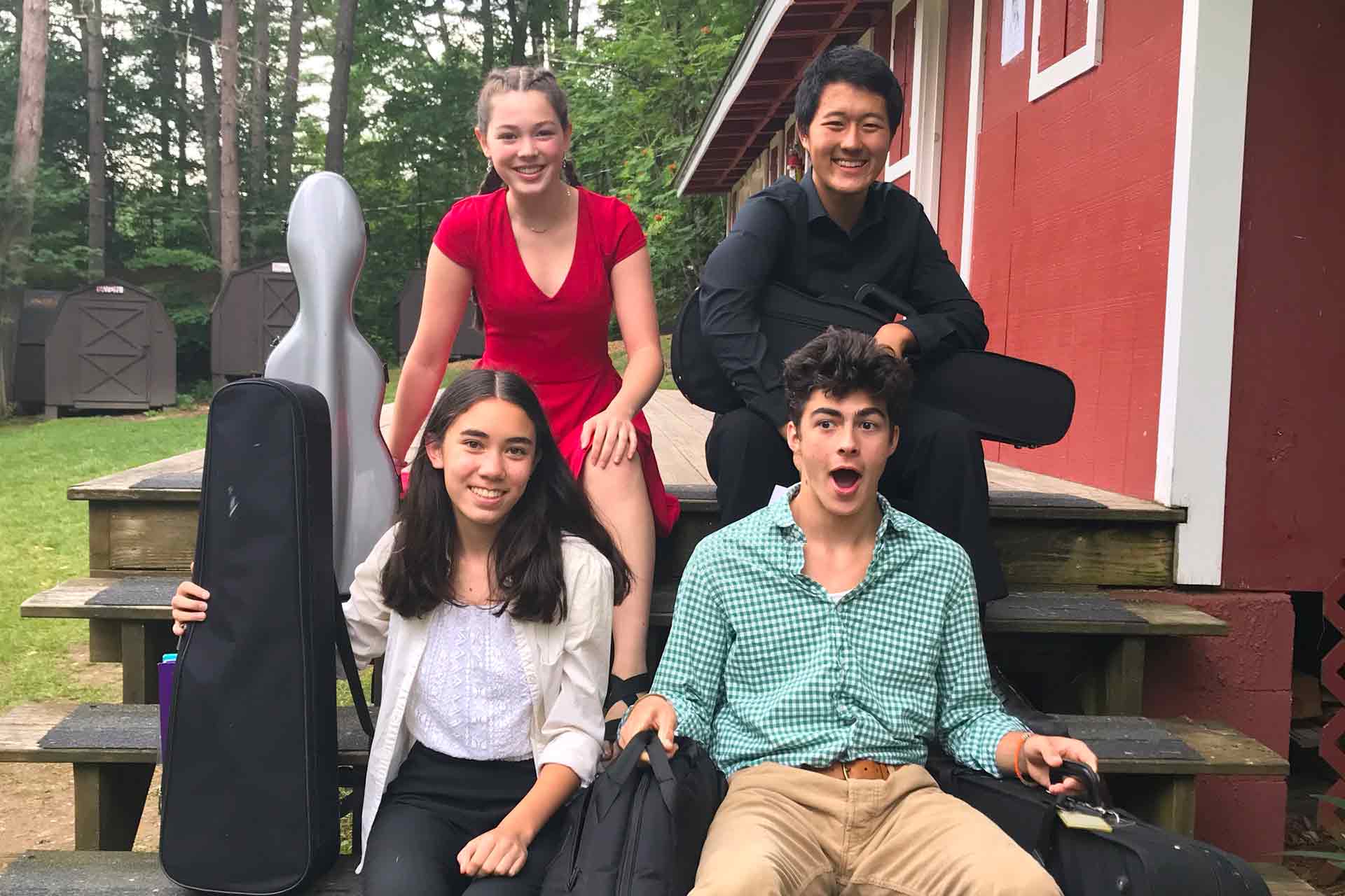 Payments and Financial Aid Four young people with musical instrument cases sit and stand on outdoor wooden steps beside a red building, smiling at the camera—perhaps discussing Financial Aid or upcoming payments for their music program. Trees and sheds are visible in the background.