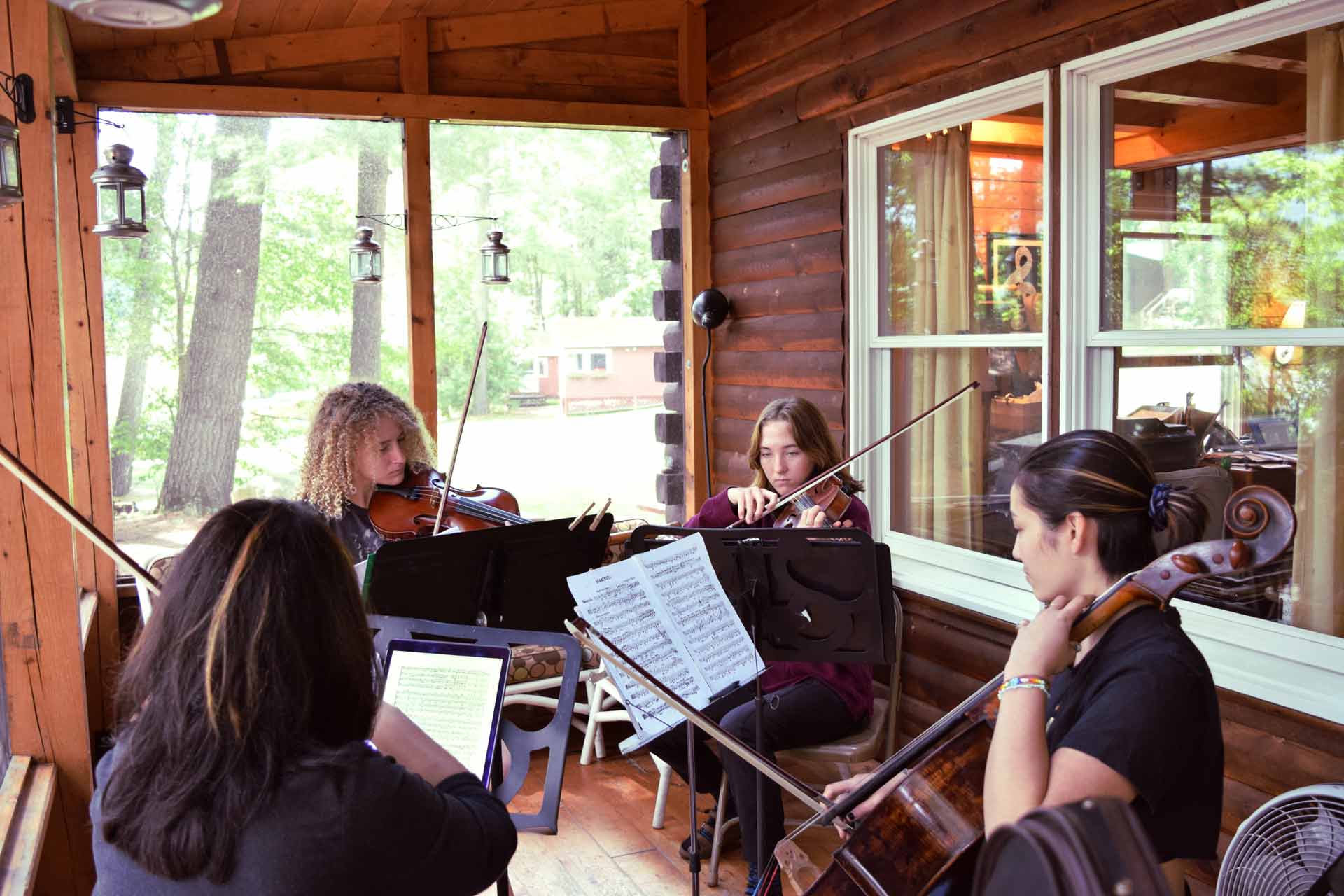 Point CounterPoint Foundation Four musicians from the Point CounterPoint Foundation play string instruments on a wooden porch, surrounded by sheet music stands, with trees and cabins visible outside.