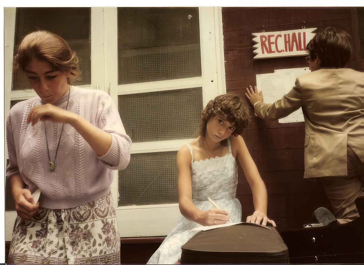 Point CounterPoint History Three young people are outside a building labeled REC. HALL; one writes notes, perhaps for a Point CounterPoint debate, one adjusts a sign on the wall, and another looks down while brushing her hand near her face—each adding their mark to history.