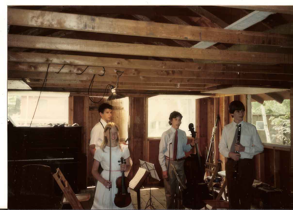 Point CounterPoint History Four young musicians in formal attire stand with their instruments—violin, cello, and clarinet—in a wooden room with exposed beams at Point CounterPoint, preparing to make history with their performance.