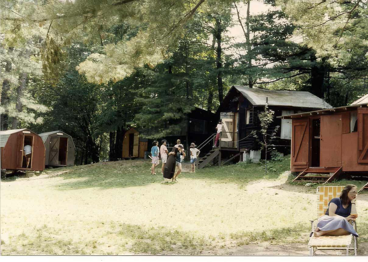 Point CounterPoint History A group of people stand near wooden cabins in a wooded camp area, while a woman—perhaps reflecting on Point CounterPoint History—sits alone on a chair in the foreground.