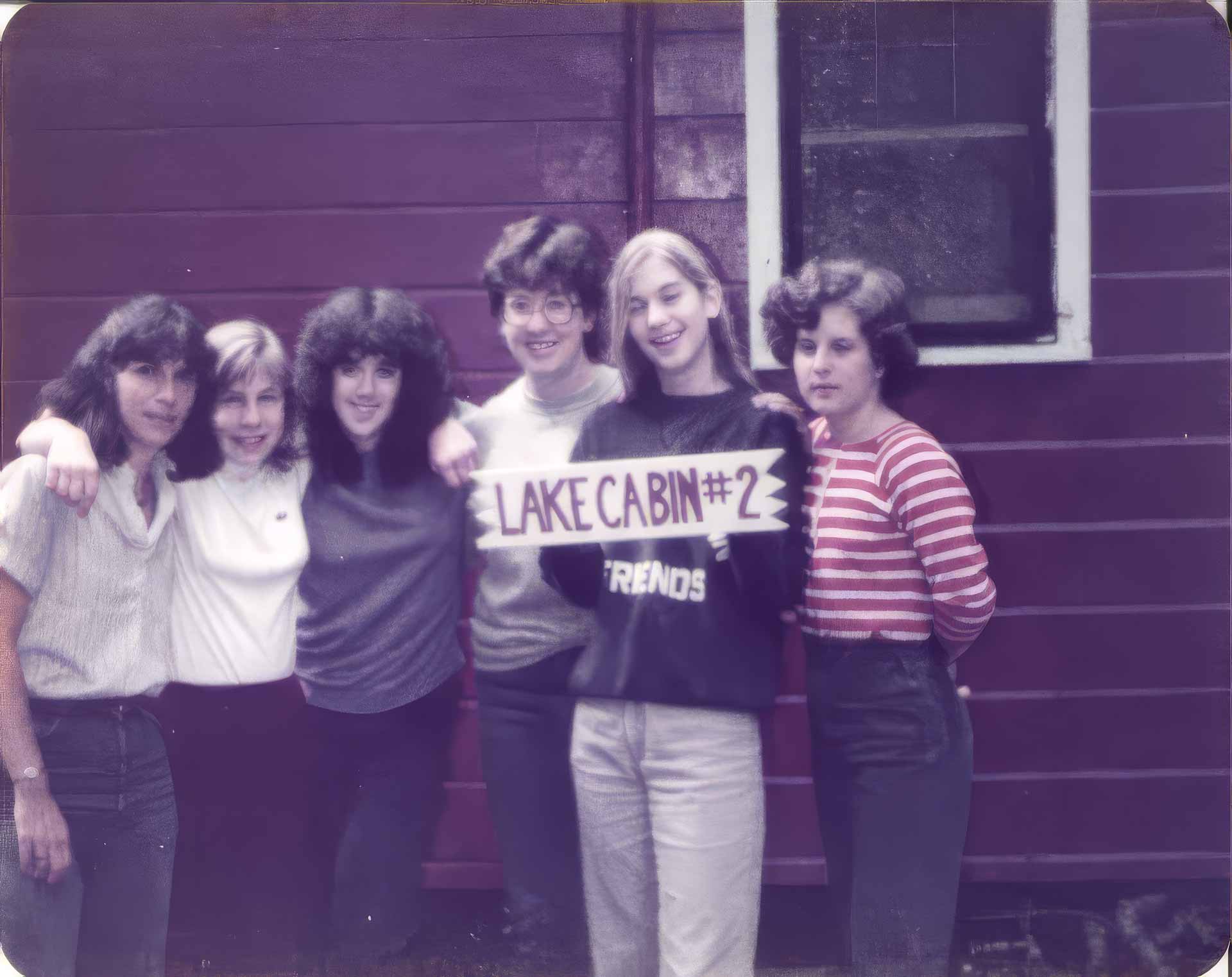 About Point CounterPoint Five women stand in front of a red wooden building, smiling at the camera. One holds a sign that reads Lake Cabin #2, capturing a cheerful moment during their Point CounterPoint retreat.