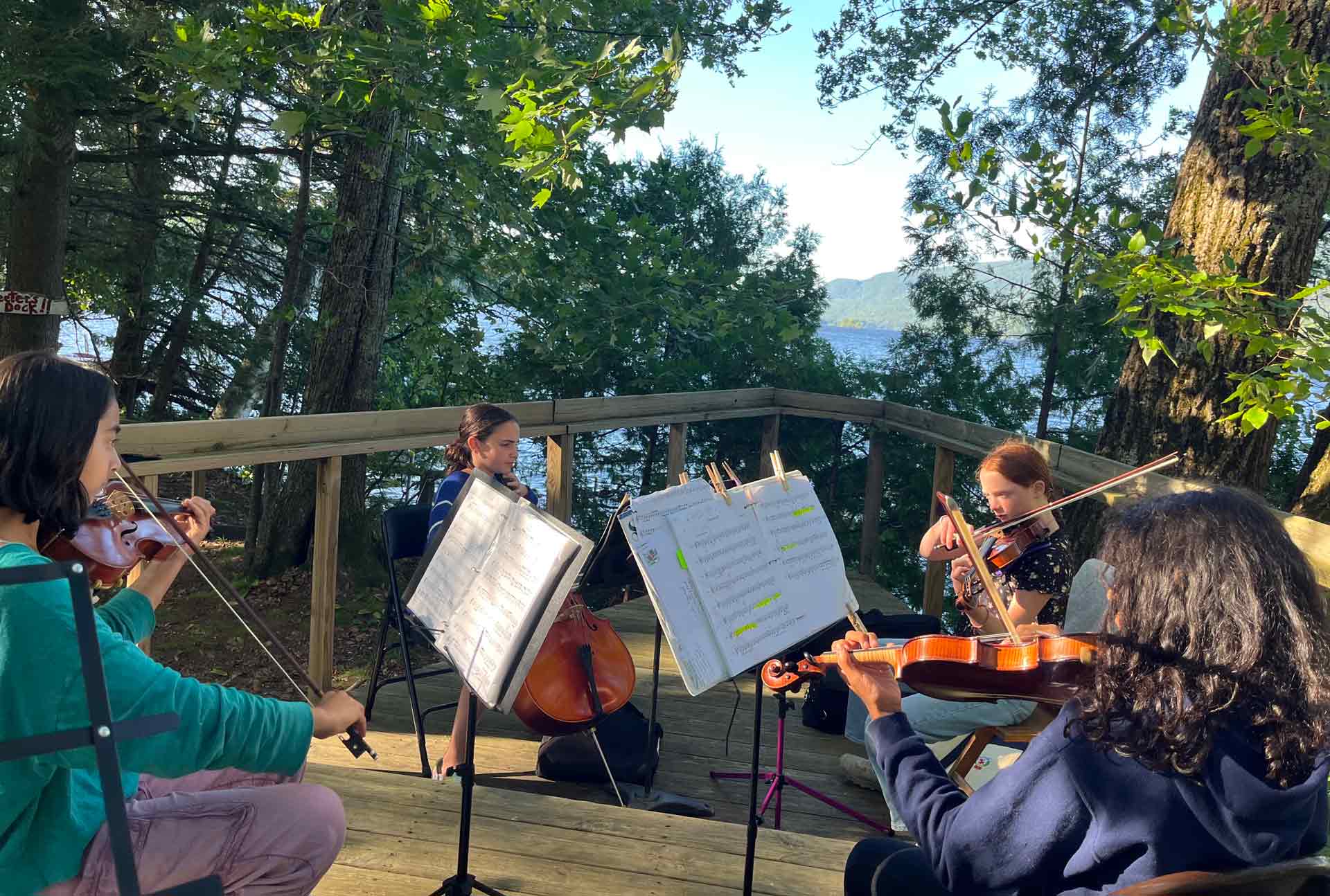 Coming to Camp! Four people play string instruments outdoors on a wooden deck surrounded by trees, with sheet music in front of them and a lake in the background—a classic Point CounterPoint moment amidst nature.