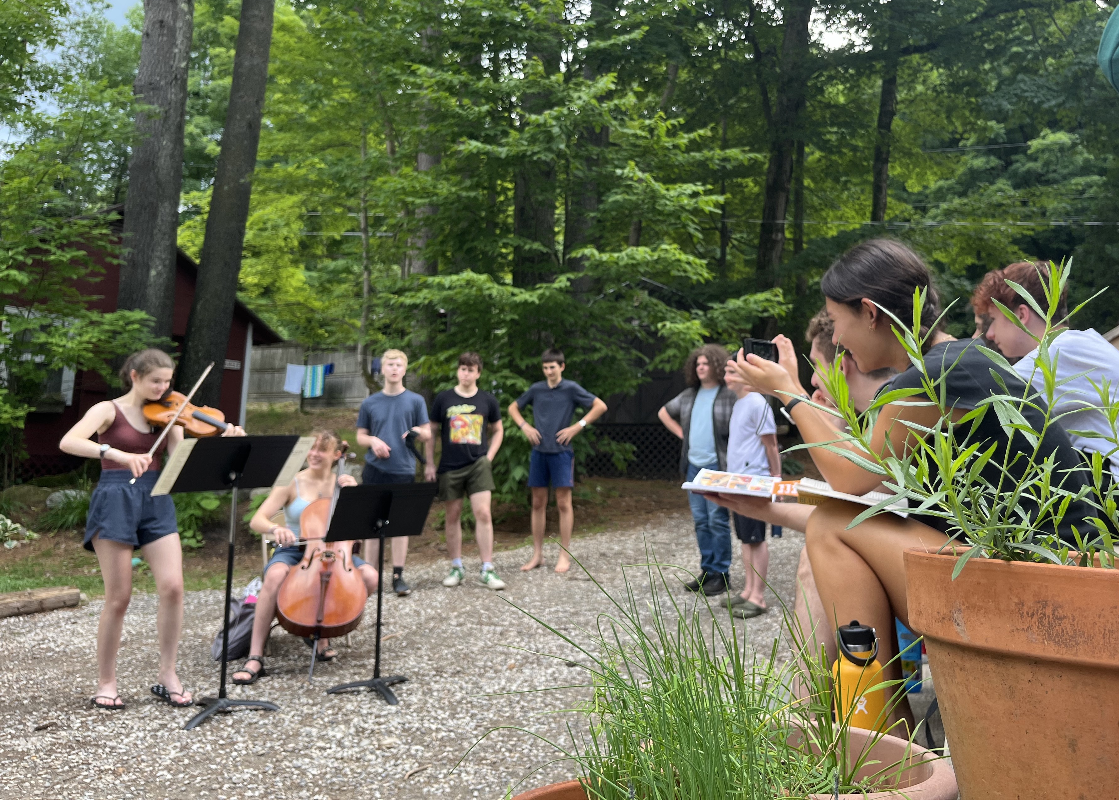 About Point CounterPoint Two young musicians perform a Point CounterPoint duet on violin and cello outdoors, as a small audience watches among trees and potted plants, some standing and others seated.