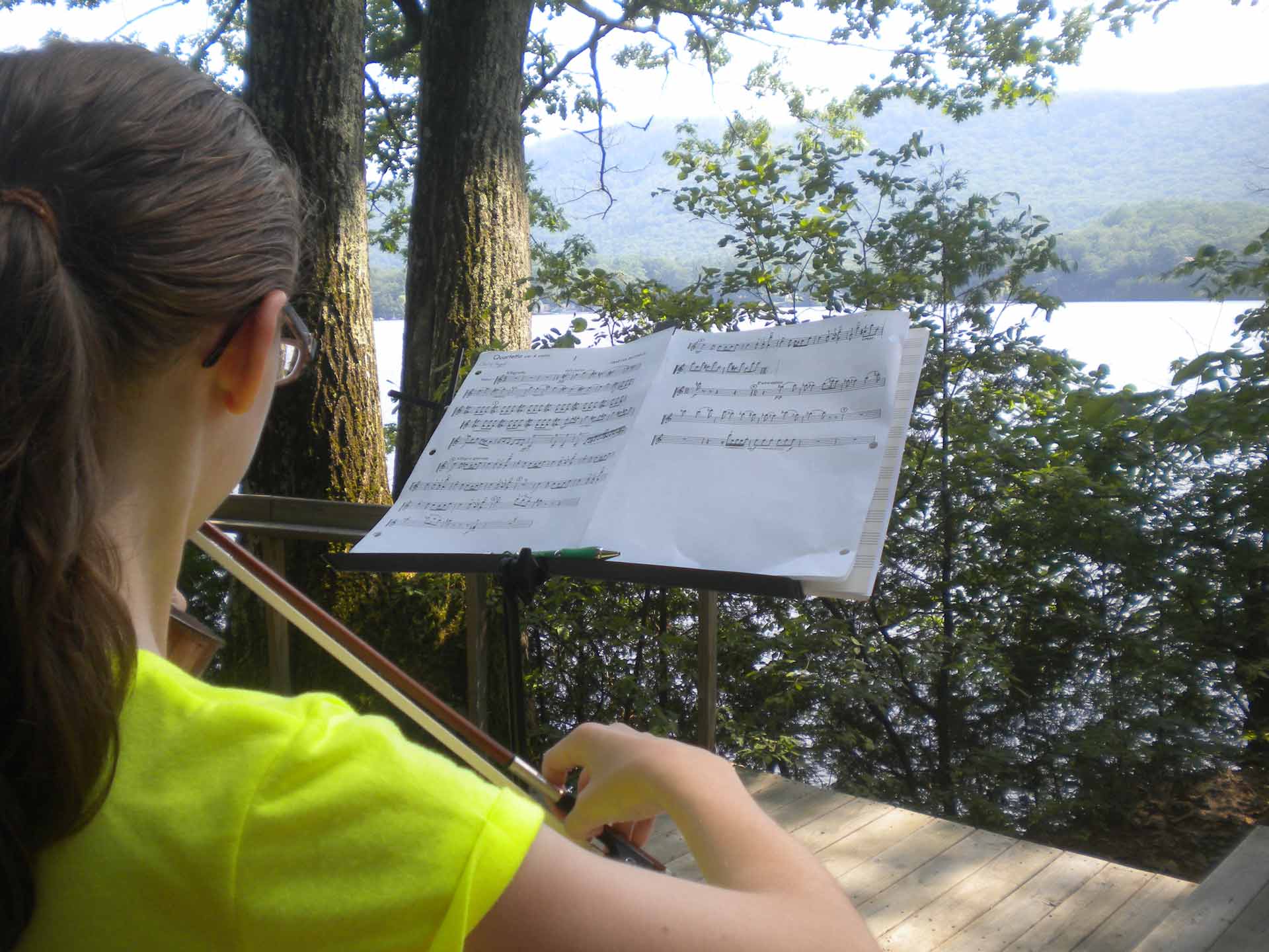 Play it Forward Campaign A person with long hair and glasses plays the violin outdoors, reading sheet music on a stand with a lake and trees in the background as part of the Play it Forward Campaign.