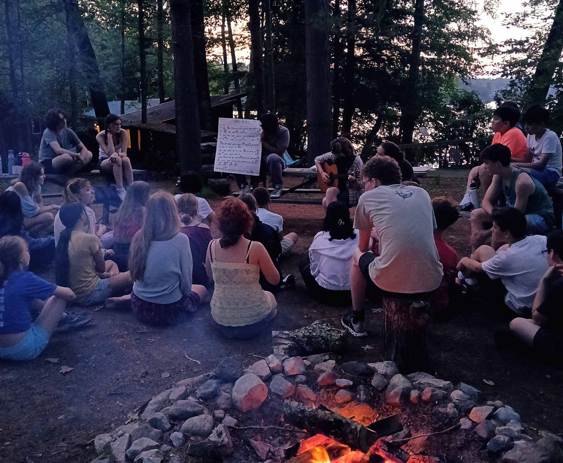 Play it Forward Campaign A group of people sit around a campfire in a wooded area, listening to someone play guitar as part of the Play it Forward Campaign; a large sheet with song lyrics is displayed nearby.