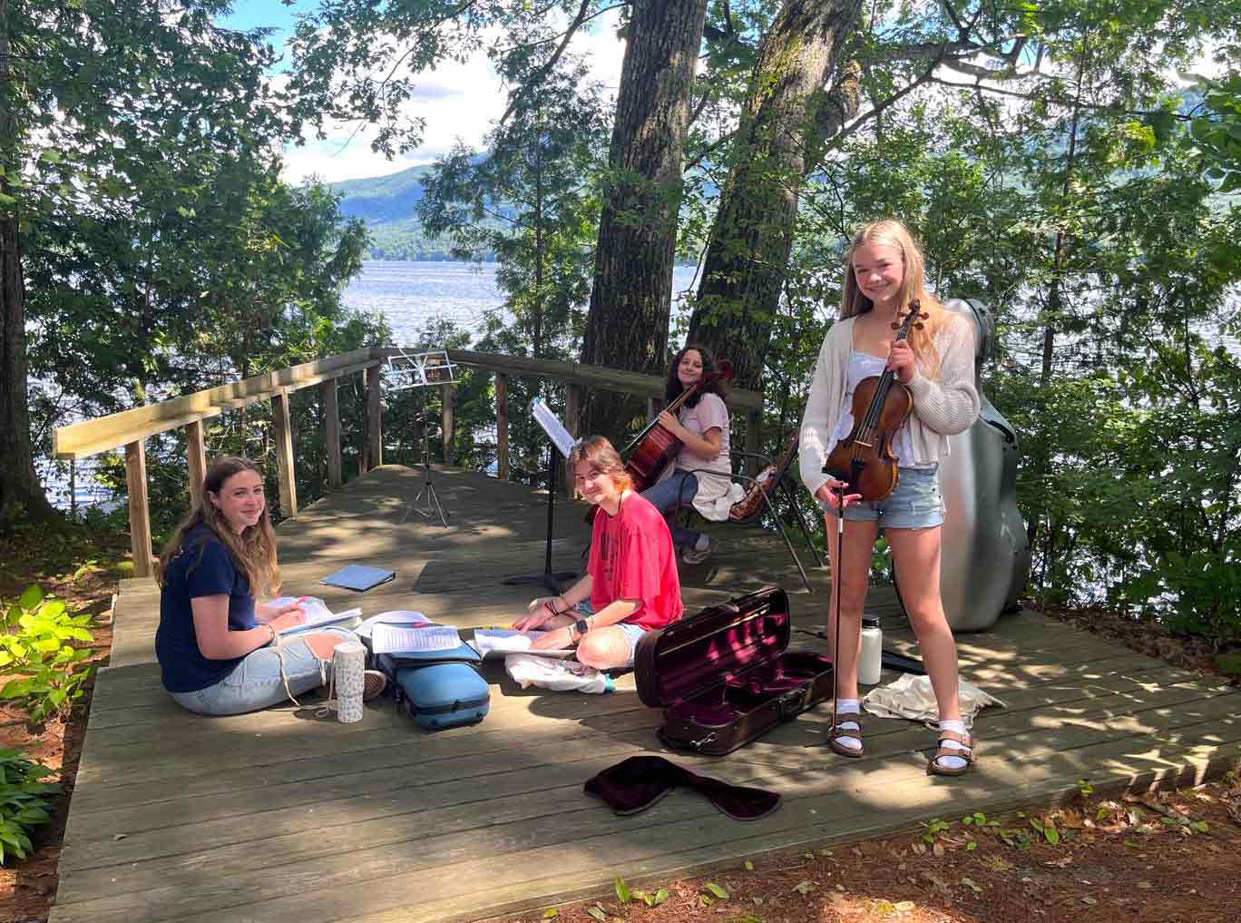 Youth Programs Four young women with string instruments and sheet music are gathered on a wooden deck by a lake, surrounded by trees in daylight.