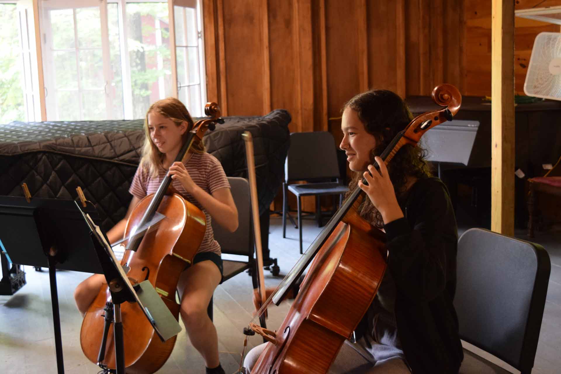 Play it Forward Campaign Two young people sit indoors playing cellos, reading sheet music on stands, with a piano and wooden walls in the background—a perfect moment of musical connection inspired by the Play it Forward Campaign.